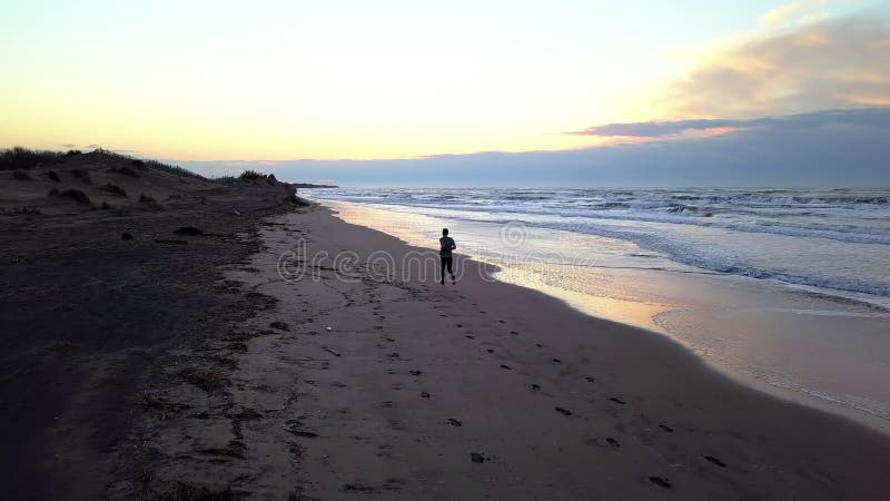 Aerial panning with drone on young attractive man jogging on the beach at sunrise. Running workout practice in fitness and healthy