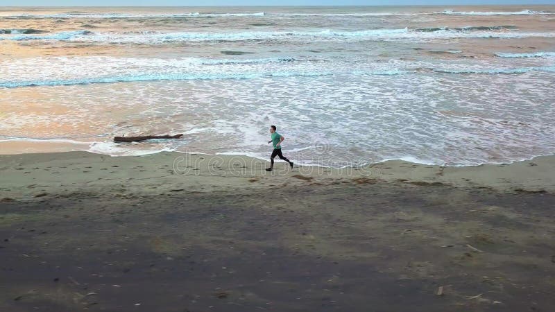 Aerial panning with drone on young attractive man jogging on the beach at sunrise. Running workout practice in fitness and healthy