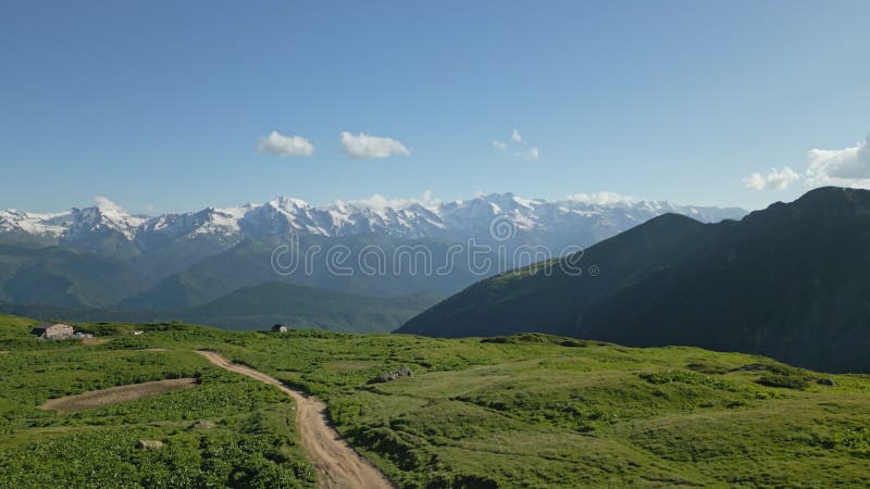 Aerial shot of a hiker walking in alpine meadows of high mountains. Green landscapes, trail paths, rugged peaks, blue stock footage