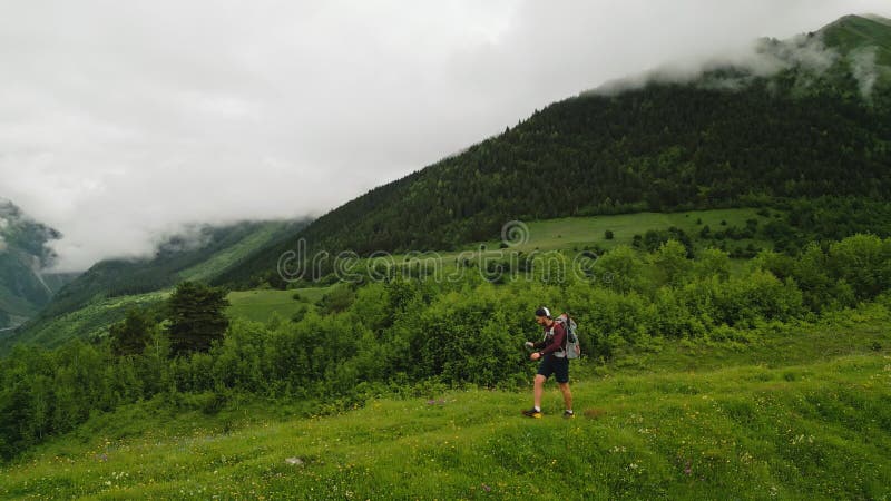 Aerial of solo backpacker hikes alpine meadows. He explores green landscapes with mountains in background. Adventurer stock video footage
