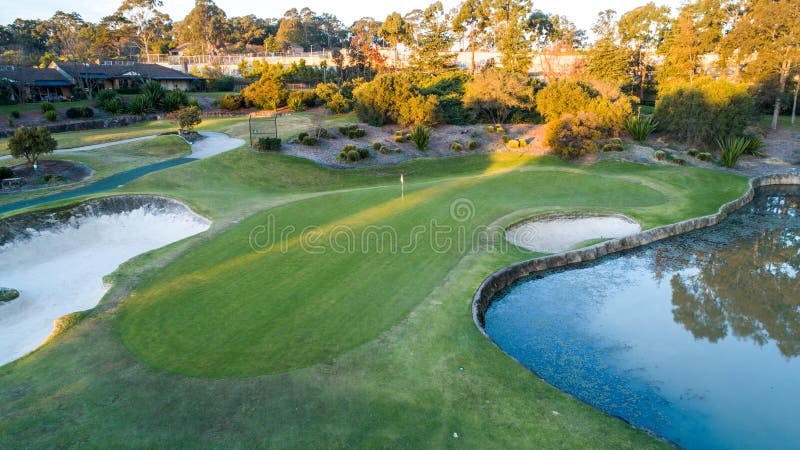 Aerial view of golf course green with flag, bunkers and dam water hazards surrounded by trees in background