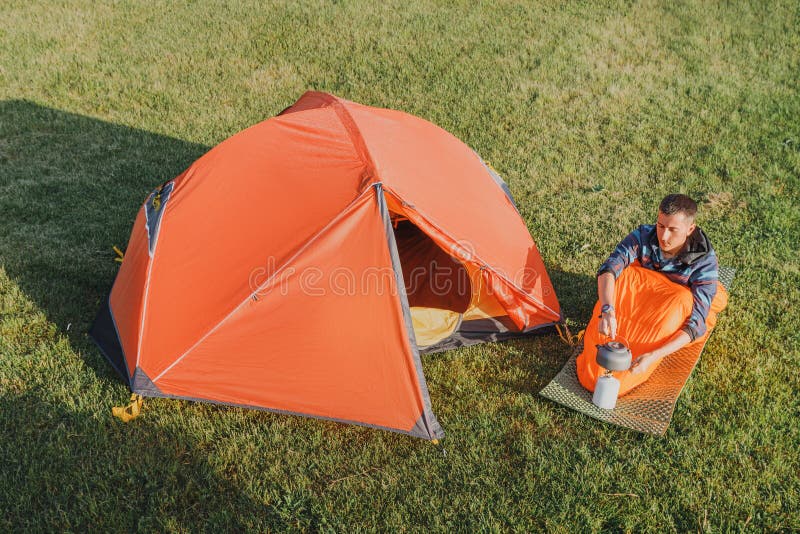 Aerial view of man boiling tea on a cooking camping gas stove or burner, near open orange tent on a meadow at early morning stock photos