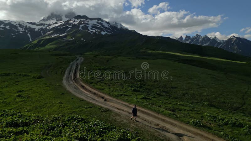 Aerial view of man hiking on alpine meadows in high mountains. Solo hiker walks on trail surrounded by green grass, snow stock video
