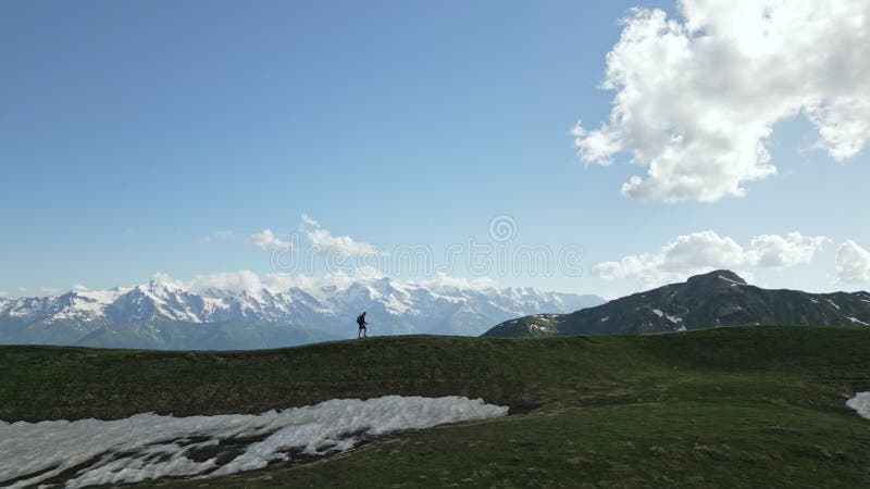 Aerial view of solo hiker walking along ridge in mountains with snowy peaks, grassy slopes. Trekking in nature stock video footage