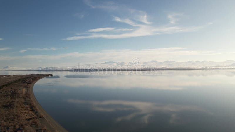 Aerial of a tranquil, reflective lake, with snowy mountain range in the background on a sunny day stock video