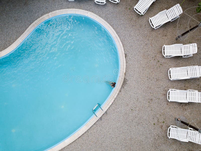 Aerial view of a young girl in black swimsuit, blue swimming pool and white sunbeds