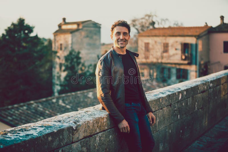 Affascinante uomo sorridente all'aperto con giacca di pelle e jeans. appoggiato su una parete bassa. vecchie case di un villaggio immagini stock