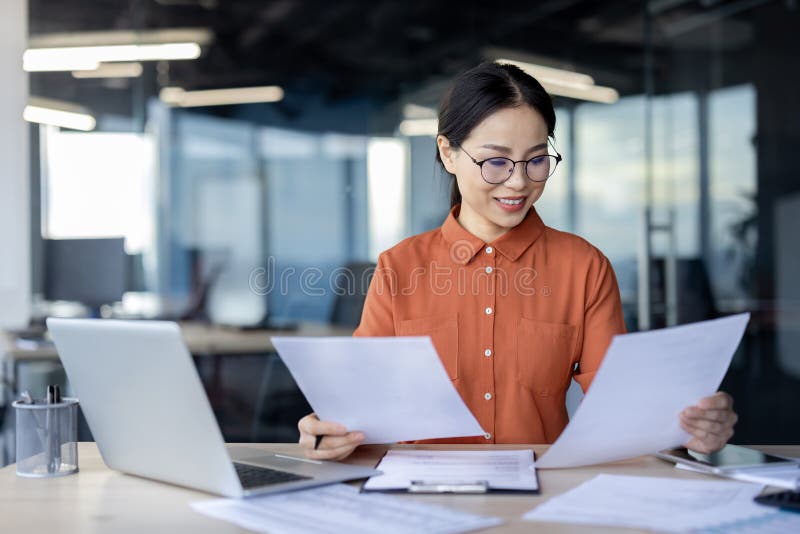 Confident Asian businesswoman happily reviews multiple documents at her desk in a contemporary office space, showcasing productivity and focus. Confident Asian businesswoman happily reviews multiple documents at her desk in a contemporary office space, showcasing productivity and focus.
