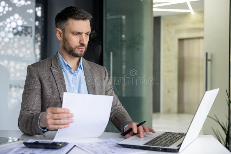 Focused professional male in a suit analyzing papers with a laptop in a contemporary office environment. Represents work, concentration, and corporate lifestyle. Focused professional male in a suit analyzing papers with a laptop in a contemporary office environment. Represents work, concentration, and corporate lifestyle.