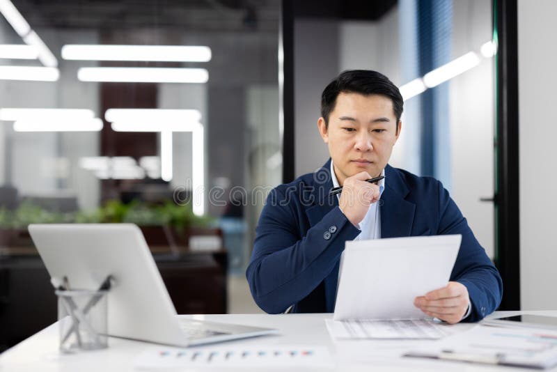 Focused Asian businessman with glasses examining paperwork at his desk in a contemporary office environment. He appears thoughtful and concentrated. Focused Asian businessman with glasses examining paperwork at his desk in a contemporary office environment. He appears thoughtful and concentrated.