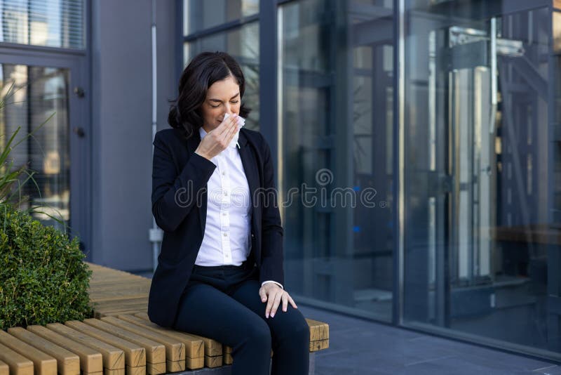 A professional businesswoman in a suit sneezing into a tissue while sitting outside a contemporary office building, capturing a moment of illness or allergy. A professional businesswoman in a suit sneezing into a tissue while sitting outside a contemporary office building, capturing a moment of illness or allergy.