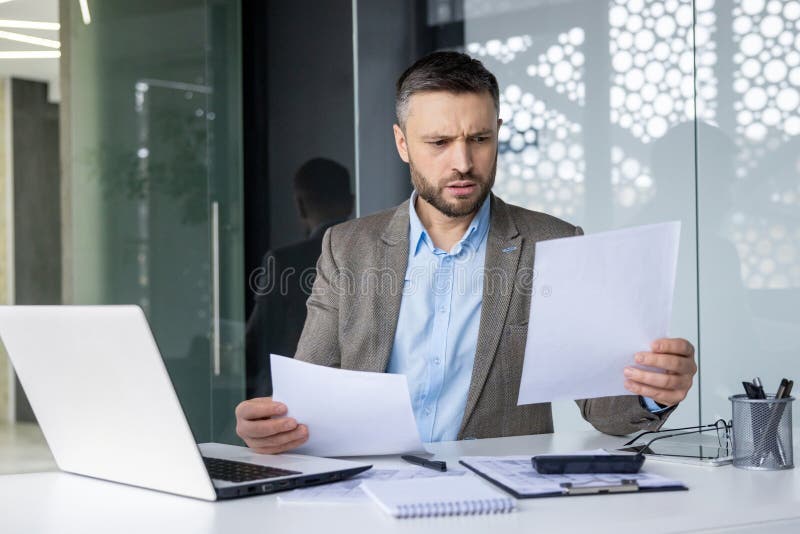 A professional businessman in a suit looks worried as he examines documents at his desk in a contemporary office setting. His laptop and office supplies are in view. A professional businessman in a suit looks worried as he examines documents at his desk in a contemporary office setting. His laptop and office supplies are in view.