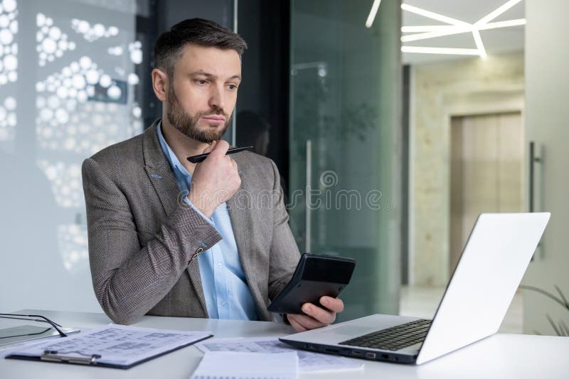 A focused businessman with a beard considers his options while reviewing financial documents using a laptop and smartphone in a contemporary office setting. A focused businessman with a beard considers his options while reviewing financial documents using a laptop and smartphone in a contemporary office setting.