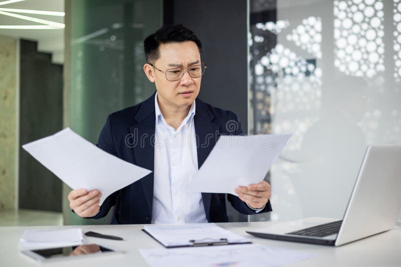 A mature Asian businessman looks perplexed while reviewing documents at his workplace, laptop open nearby in a stylish office setting. A mature Asian businessman looks perplexed while reviewing documents at his workplace, laptop open nearby in a stylish office setting.