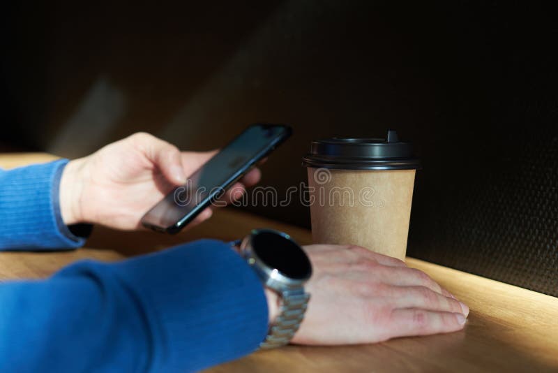 Affärsmannen dricker kaffe i ett kafé, närbild rymmer ett disponibelt pappers- exponeringsglas, genom att använda smartphonen, me royaltyfri foto Affärsmannen dricker kaffe i ett kafé, närbild rymmer ett disponibelt pappers- exponeringsglas, genom att använda smartphonen, me royaltyfri foto