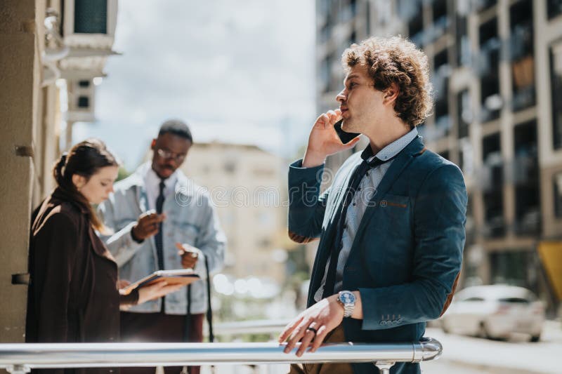 Three professionals engage in a business discussion on a sunlit balcony, with a man making a phone call and his colleagues reviewing documents. Three professionals engage in a business discussion on a sunlit balcony, with a man making a phone call and his colleagues reviewing documents.