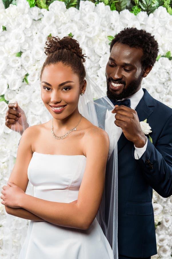 African american bridegroom touching white veil and smiling near bride and flowers. Cheerful african american bridegroom touching white veil and smiling near royalty free stock images African american bridegroom touching white veil and smiling near bride and flowers. Cheerful african american bridegroom touching white veil and smiling near royalty free stock images