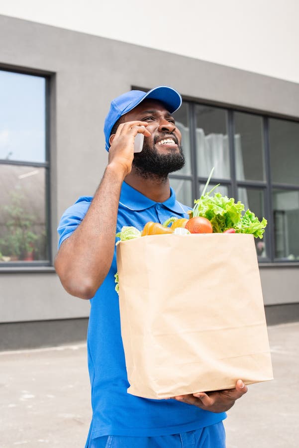 african american delivery man holding paper bag with vegetables and talking