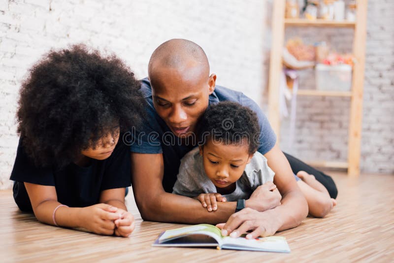 African American father reading a fairy tale fable story for kids at home. Happy family lying on the floor indoors