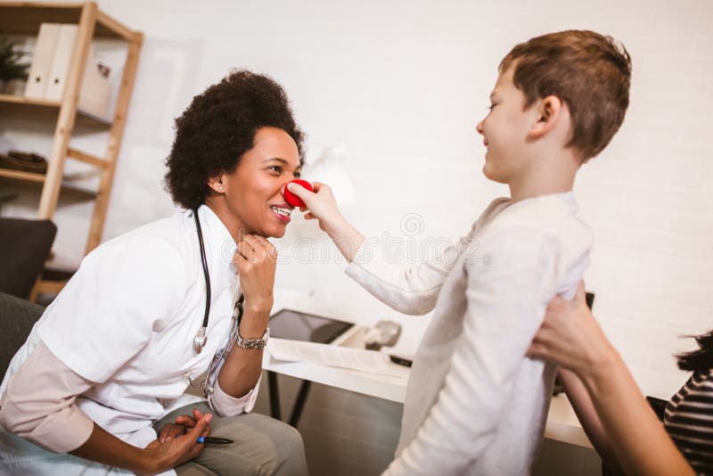 African American female pediatrician with stethoscope and clown nose talking to boy