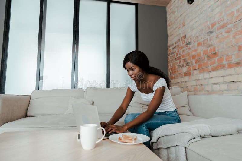 African american freelancer using laptop at coffee table with cup of tea and plate with toasts in living room