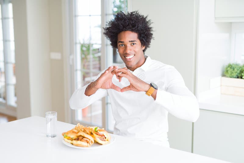 African American hungry man eating hamburger for lunch smiling in love showing heart symbol and shape with hands