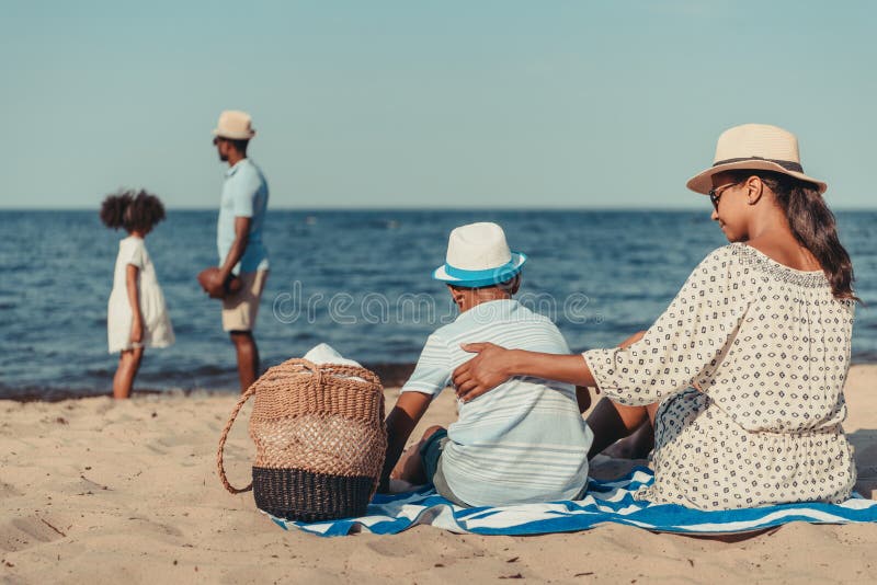 african american mother and son sitting on sand while father and daughter playing