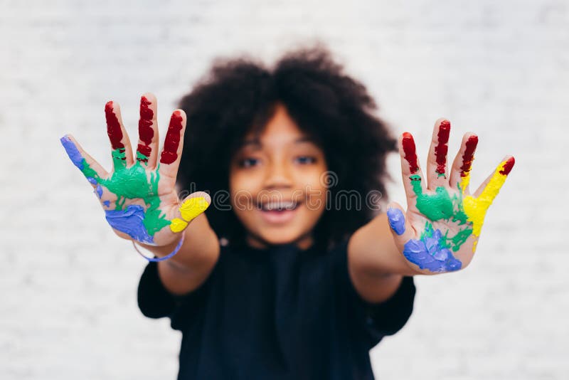 African American playful and creative kid getting hands dirty with many colors - in white brick background.