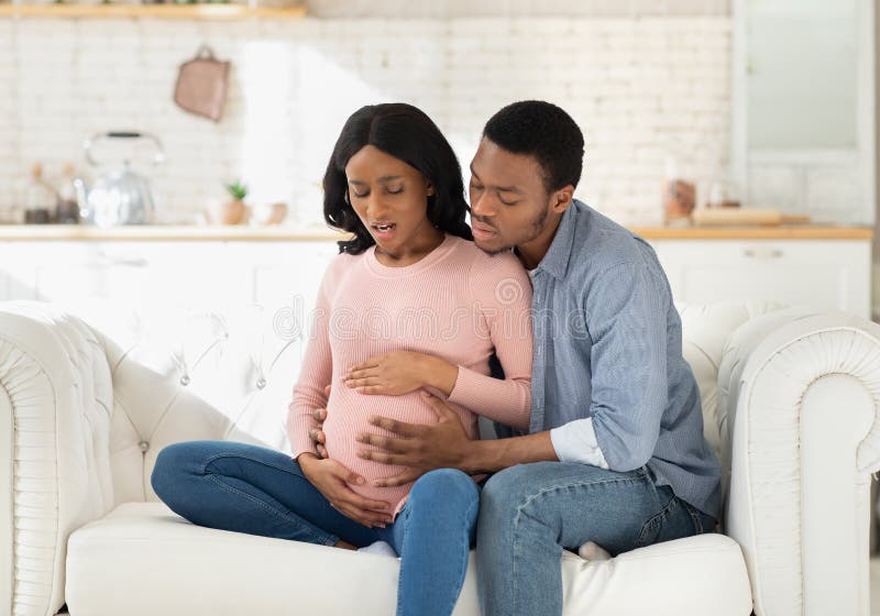 African American pregnant woman suffering from pain, having birth contractions while worried husband helping her at home