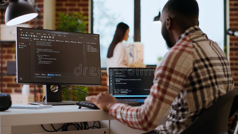 African american IT professional types programming code on dual monitors stock images
