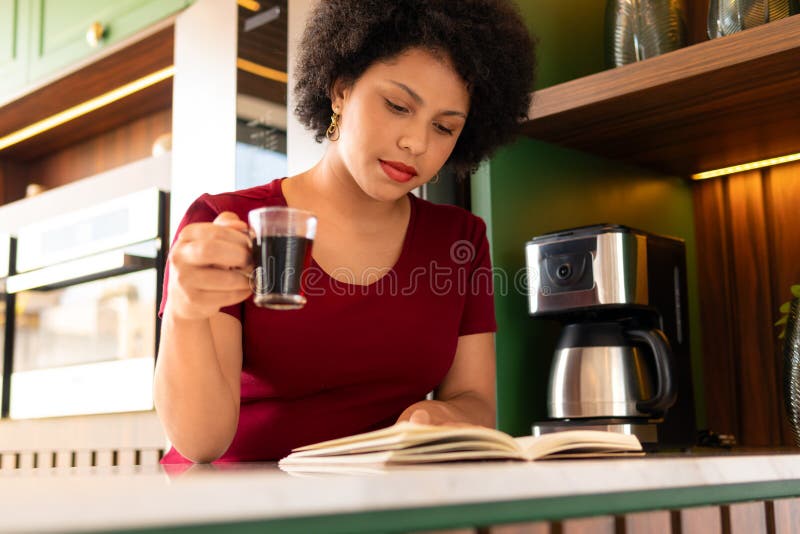 Woman Reading Book And Drinking Coffee In Kitchen Counter Stock Image