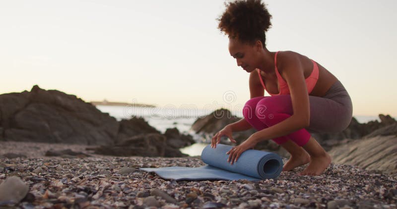 African american woman rolling her yoga mat on the rocks near the sea during sunset