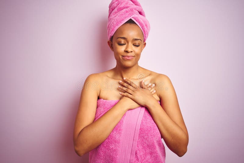 African american woman wearing shower towel after bath over pink isolated background smiling with hands on chest with closed eyes