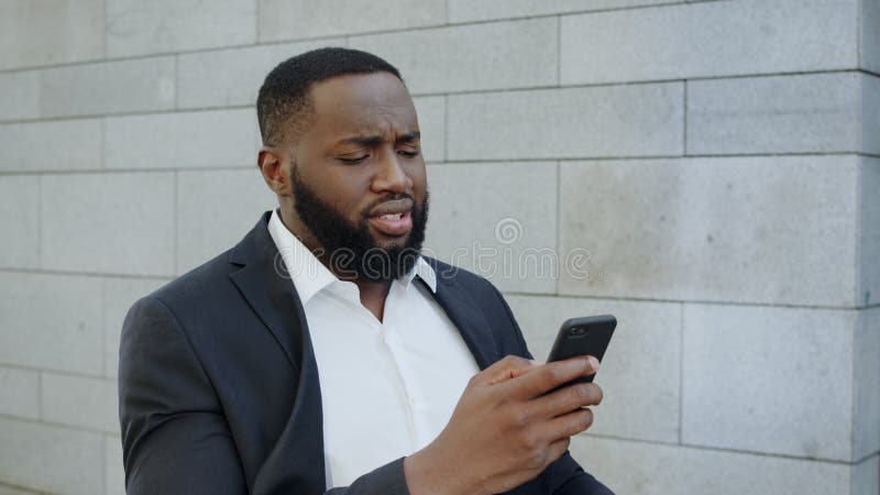 African businessman getting bad news on mobile phone in modern city. Upset male employee using smartphone outside office. Sad african american business man walking on street in slow motion. African businessman getting bad news on mobile phone in modern city. Upset male employee using smartphone outside office. Sad african american business man walking on street in slow motion