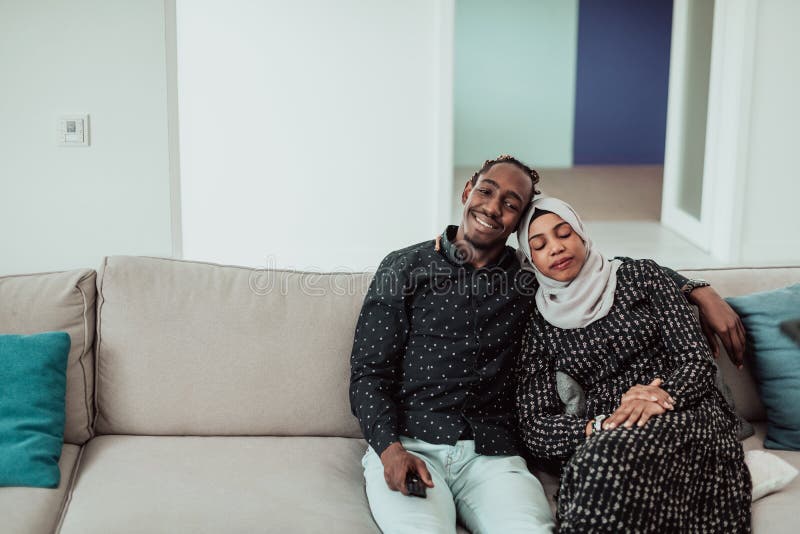 African Couple Sitting On Sofa Watching TV Together. Woman Wearing Islamic Hijab Clothes