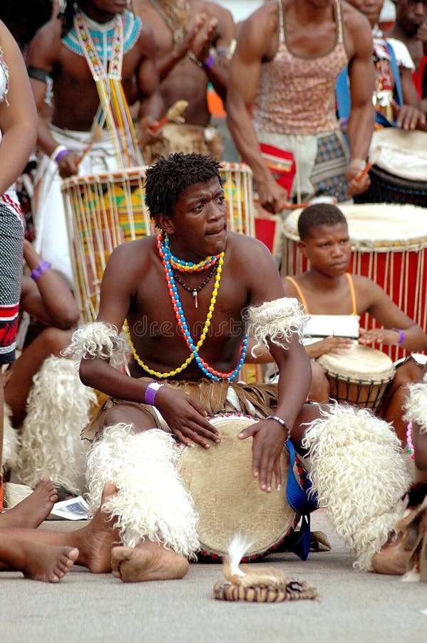 A black African musician playing a traditional drum in front of a group of dancers on the street outdoors. A black African musician playing a traditional drum in front of a group of dancers on the street outdoors