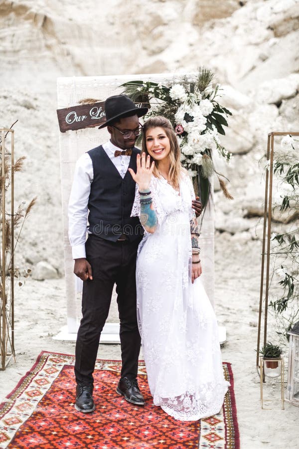African groom and Caucasian bride embracing each other in a boho style before the wedding arch from fresh flowers. Bride royalty free stock photo African groom and Caucasian bride embracing each other in a boho style before the wedding arch from fresh flowers. Bride royalty free stock photo