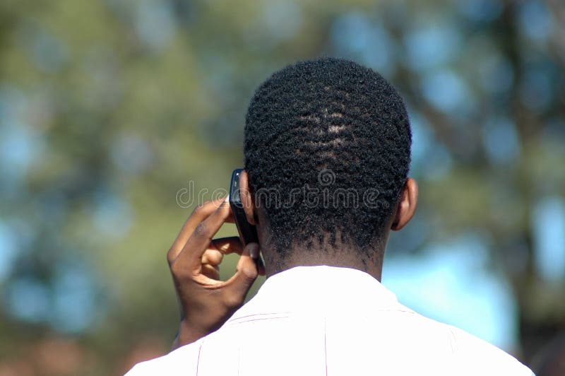 An African young business man at work holding his mobile phone in his black hand and phoning in a park outdoors in South Africa. An African young business man at work holding his mobile phone in his black hand and phoning in a park outdoors in South Africa