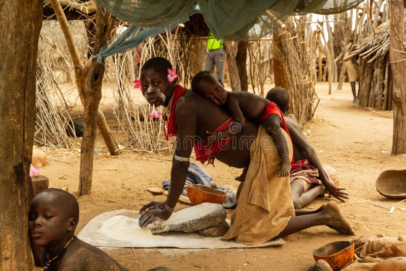 An African woman grinds wheat by rubbing one stone over another while carrying a baby on her back.