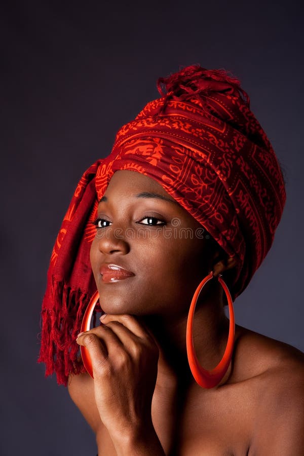Bella di etnia Afro-Americana donna che indossa un tradizionale tribale rosso arancione sciarpa in testa e big orange orecchini a cerchio, per appoggiare il mento con la mano, isolato.