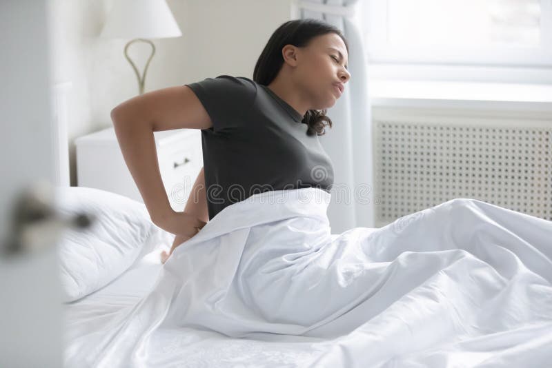 African mixed-race woman waking up sitting on bed feel low back pain after sleep caused by uncomfortable mattress, wrong position during night rest, premenstrual syndrome, violation of posture concept. African mixed-race woman waking up sitting on bed feel low back pain after sleep caused by uncomfortable mattress, wrong position during night rest, premenstrual syndrome, violation of posture concept