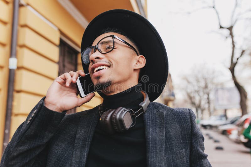 Africano disgraziato, con capelli corti, al telefono con sorriso. Foto all'aperto di un giovane nero entusiasta all'interno fotografia stock