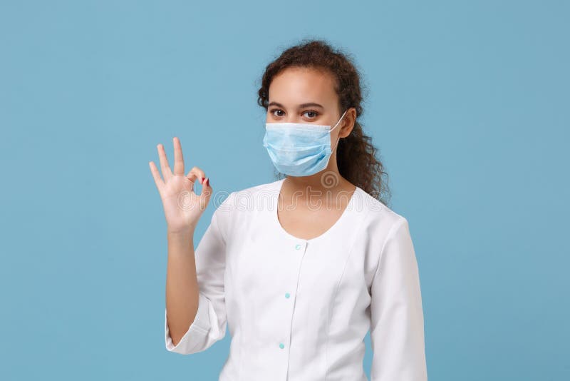 African american doctor woman isolated on blue background. Female doctor in white medical gown, sterile face mask showing OK gesture. Healthcare personnel health medicine concept. Mock up copy space. African american doctor woman isolated on blue background. Female doctor in white medical gown, sterile face mask showing OK gesture. Healthcare personnel health medicine concept. Mock up copy space