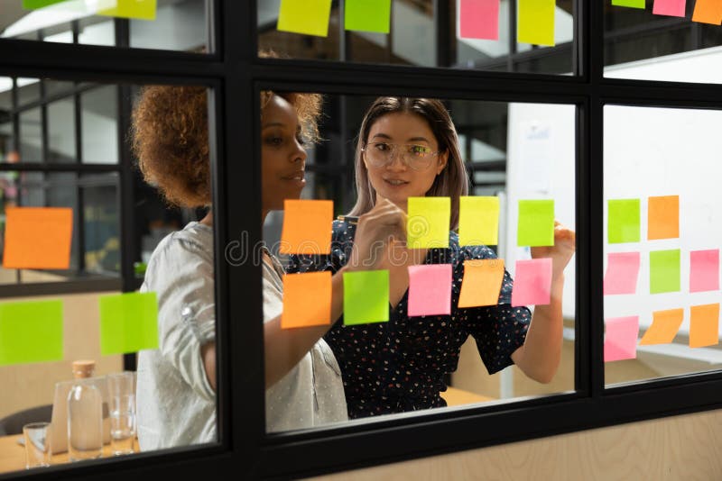 Concentrated african american young female scrum master working together with smart vietnamese businesswoman colleague, putting notes on sticky paper at window glass agile kanban board at office. Concentrated african american young female scrum master working together with smart vietnamese businesswoman colleague, putting notes on sticky paper at window glass agile kanban board at office.