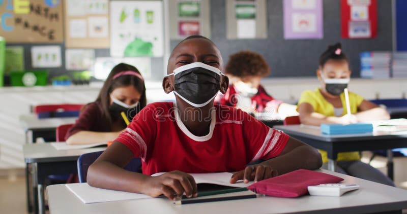 Afrikaanse amerikaanse jongen die een gezichtsmasker draagt terwijl hij op zijn bureau zit in de klas op school