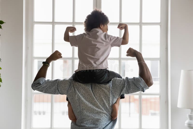 Back side view young african american father holding on shoulders little preschool funny kid son, showing biceps. Fit strong different generations family enjoying domestic activity, healthcare concept. Back side view young african american father holding on shoulders little preschool funny kid son, showing biceps. Fit strong different generations family enjoying domestic activity, healthcare concept.