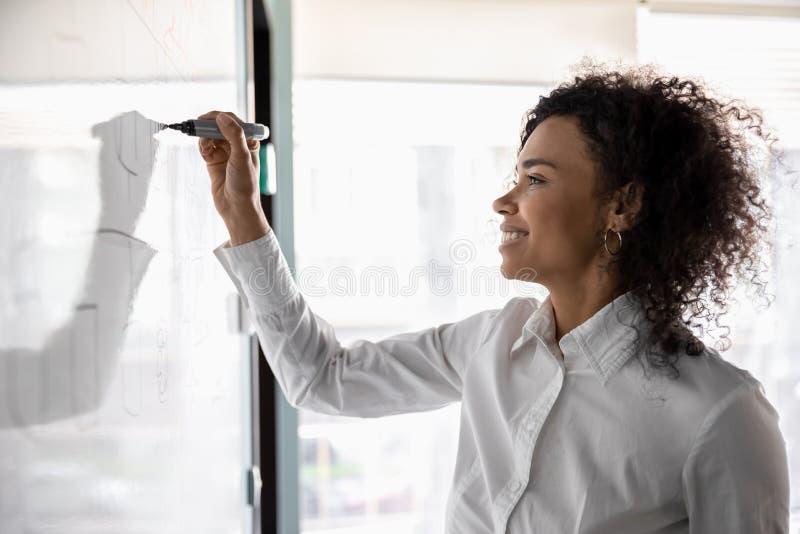 Smiling young african American businesswoman busy writing on white board developing company financial business plan, millennial biracial woman brainstorm involved in creative thinking use flip chart. Smiling young african American businesswoman busy writing on white board developing company financial business plan, millennial biracial woman brainstorm involved in creative thinking use flip chart