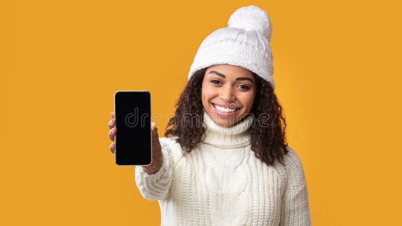 Modern Technology Concept. Smiling african american woman in winter hat holding cellphone with empty black screen over orange studio background. Happy lady showing gadget display for mock up. Modern Technology Concept. Smiling african american woman in winter hat holding cellphone with empty black screen over orange studio background. Happy lady showing gadget display for mock up