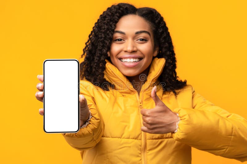 African American Woman Showing Phone Empty Screen To Camera Gesturing Thumbs Up Approving Smartphone Standing On Yellow Background, Wearing Winter Jacket. Cellphone Display Mockup. Shallow Depth. African American Woman Showing Phone Empty Screen To Camera Gesturing Thumbs Up Approving Smartphone Standing On Yellow Background, Wearing Winter Jacket. Cellphone Display Mockup. Shallow Depth