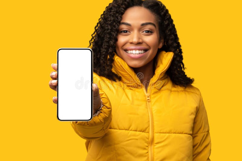 Smiling African Girl Showing Mobile Phone Screen Posing On Yellow Studio Background, Wearing Winter Clothes. Smiling African Girl Showing Mobile Phone Screen Posing On Yellow Studio Background, Wearing Winter Clothes